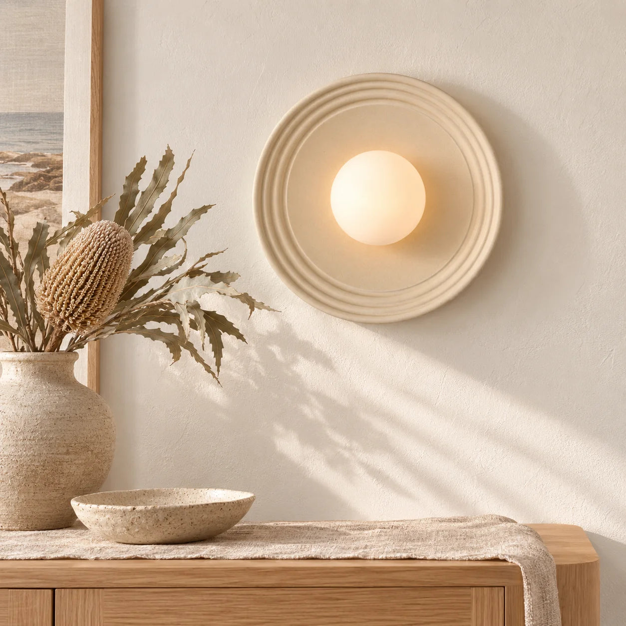 A warm, modern wall sconce with a circular ribbed beige frame and a glowing frosted globe, mounted on a textured cream wall above a wooden console table with a vase of dried grasses.