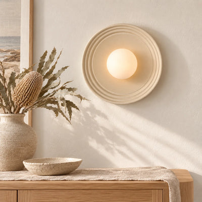 A warm, modern wall sconce with a circular ribbed beige frame and a glowing frosted globe, mounted on a textured cream wall above a wooden console table with a vase of dried grasses.